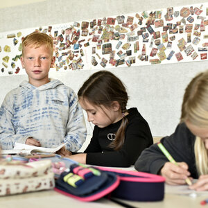Ein Schüler der Grundschule steht am Tisch und schaut ins Bild, um ihn herum arbeiten Schülerinnen und eine Lehrerin sitzt nehmen ihm